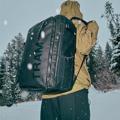 Person wearing a large black backpack with 'TRAMLINE' branding in a snowy forest setting